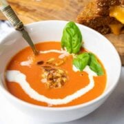 A bowl of creamy tomato soup, drizzled with cream, garnished with pine nuts and fresh basil.