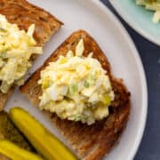 A plate with toasted bread with egg salad, pickles and potato chips.
