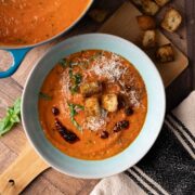 A bowl of cherry tomato soup garnished with croutons, grated cheese, chili oil and fresh basil.