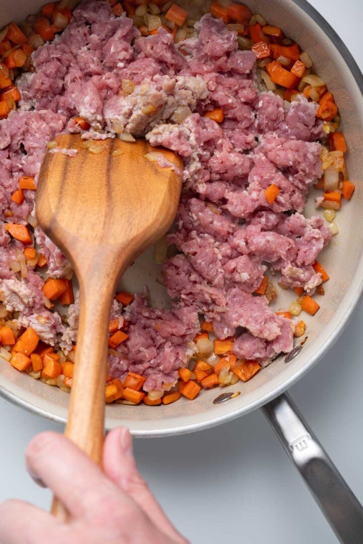 Raw ground pork in a skillet with cooked carrots and onions.