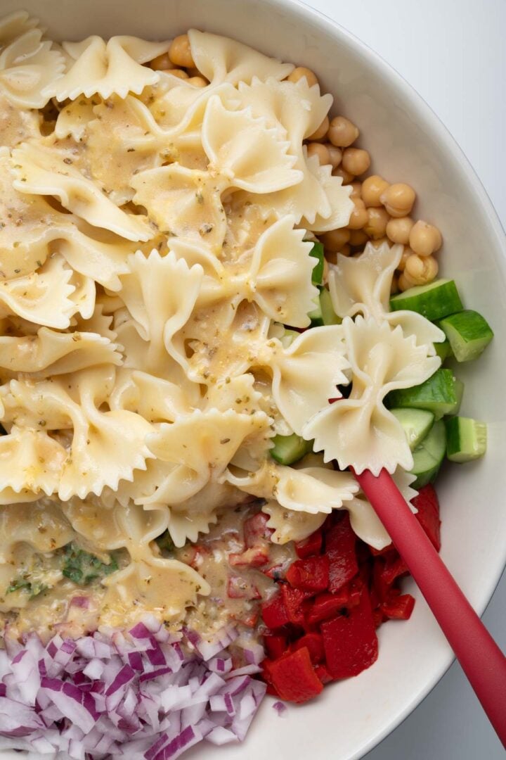 Mixing onions, red pepper, farfalle, dressing, cucumbers and chickpeas in a bowl.
